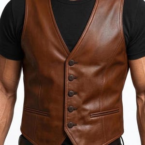 Könnte beinhalten: Eine braune Lederweste mit V-Ausschnitt und vier Knöpfen. Die Weste hat zwei Vordertaschen und wird über einem schwarzen T-Shirt getragen. Das Leder hat ein leichtes Used-Aussehen.