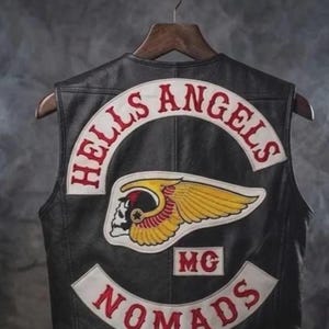 May include: Black leather vest with the "HELLS ANGELS" patch in red and white arc, and "NOMADS" patch below. Features a skull with wings emblem and "MG" patch. The vest is hanging on a wooden hanger.