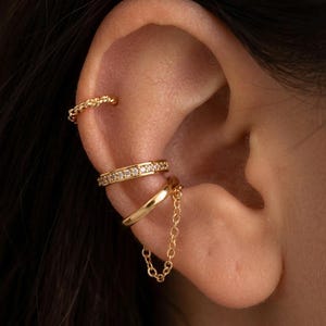May include: Gold ear cuff set featuring three different designs. One cuff has a rope-like texture, another is adorned with small, clear stones, and the third connects to a chain. The jewelry is displayed on an ear.