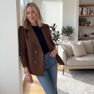 May include: A double-breasted brown blazer featuring ribbed knit sleeves, paired with a black top and blue denim jeans. The blazer has a notched lapel and two visible buttons on the front, combining a structured bodice with textured sleeves.