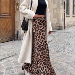 Op de afbeelding: Een crèmekleurige jas met lange mouwen gedragen over een zwarte crop top en een enkellange rok met luipaardprint. De outfit bevat een zwarte schoudertas met een dunne riem en witte enkellaarsjes. De rok heeft een hoge taille met een dunne zwarte riem.