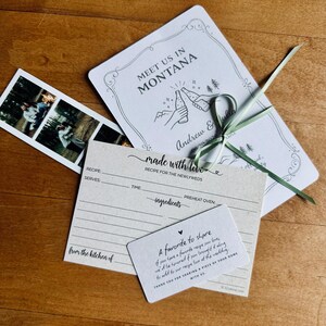 May include: A collection of wedding stationery, including an invitation with the text "Meet Us In Montana", recipe cards, and a photo strip. The invitation features a drawing of two people toasting with bottles. Tied with a green ribbon.