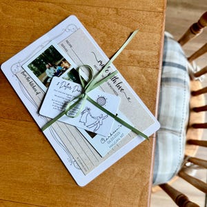 May include: A wedding recipe card set tied with a green ribbon. The set includes a recipe card, a photo, and a tag with the text "made with love" and "Clarkson's 06.20.2026 HAUGAN MT". The set is on a wooden table.