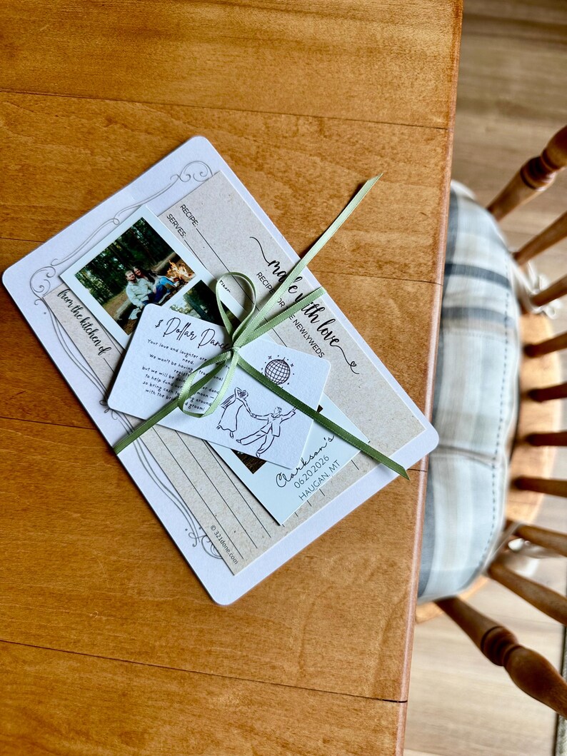 May include: A wedding recipe card set tied with a green ribbon. The set includes a recipe card, a photo, and a tag with the text "made with love" and "Clarkson's 06.20.2026 HAUGAN MT". The set is on a wooden table.