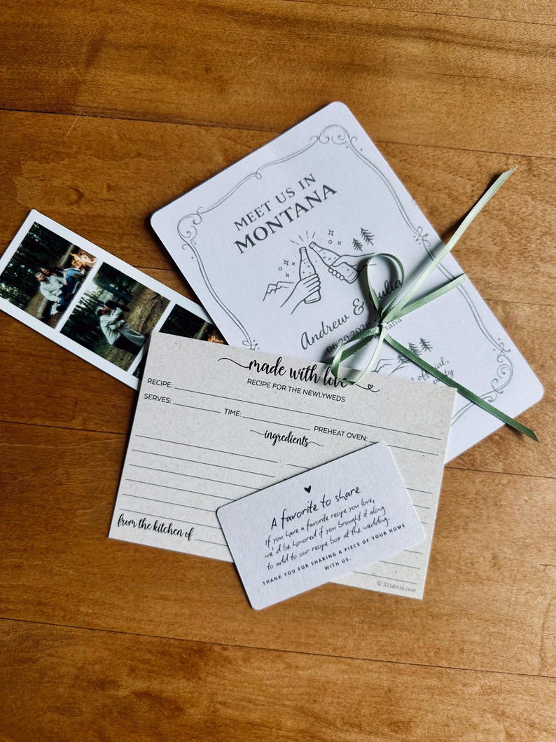 May include: Wedding stationery set featuring an invitation with the text "Meet Us In Montana", recipe cards, and a photo strip. The invitation has a drawing of two people toasting with bottles. Tied with a green ribbon.