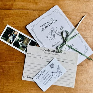 May include: Wedding stationery set on a wooden surface. Includes a photo strip, invitation with "Meet Us In Montana" text, recipe card, and a card with dancing figures and text "$ Dollar Dance $."