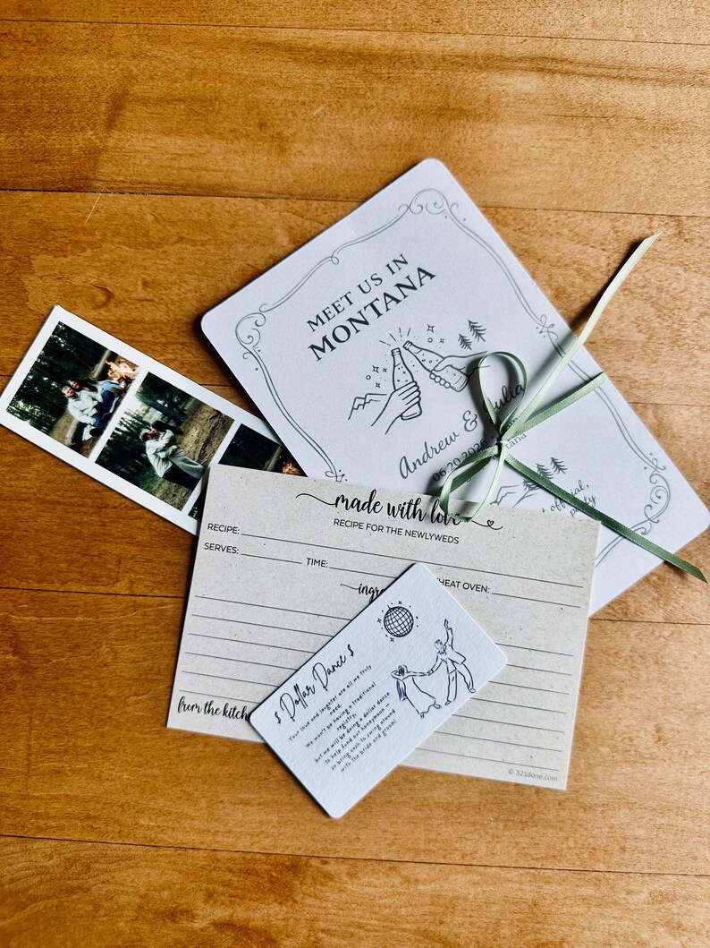 May include: Wedding stationery set on a wooden surface. Includes a photo strip, invitation with "Meet Us In Montana" text, recipe card, and a card with dancing figures and text "$ Dollar Dance $."