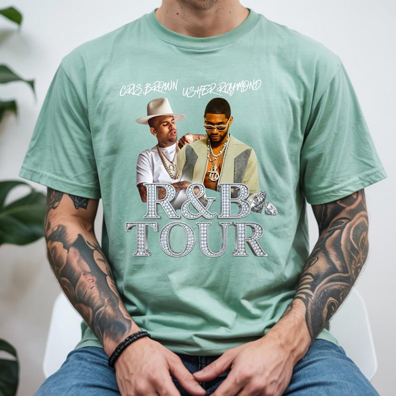 May include: A light green t-shirt featuring a graphic of Chris Brown and Usher Raymond. The text "R&B TOUR" is displayed in a sparkling, diamond-like font. The names "Chris Brown" and "Usher Raymond" are printed above the graphic.