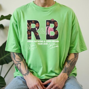May include: A light green t-shirt featuring the letters "R&B" with images of Usher Raymond and Chris Brown. The text "USHER RAYMOND & CHRIS BROWN TOUR 2026" is printed below the letters, along with two signatures.