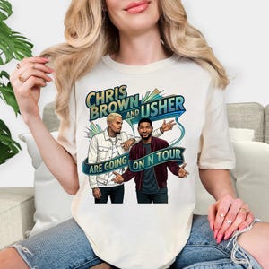 May include: Cream-colored t-shirt featuring a graphic of Chris Brown and Usher. The design includes the text "CHRIS BROWN and USHER ARE GOING ON N TOUR" in a stylized font with blue and green accents. The illustration depicts the two artists.