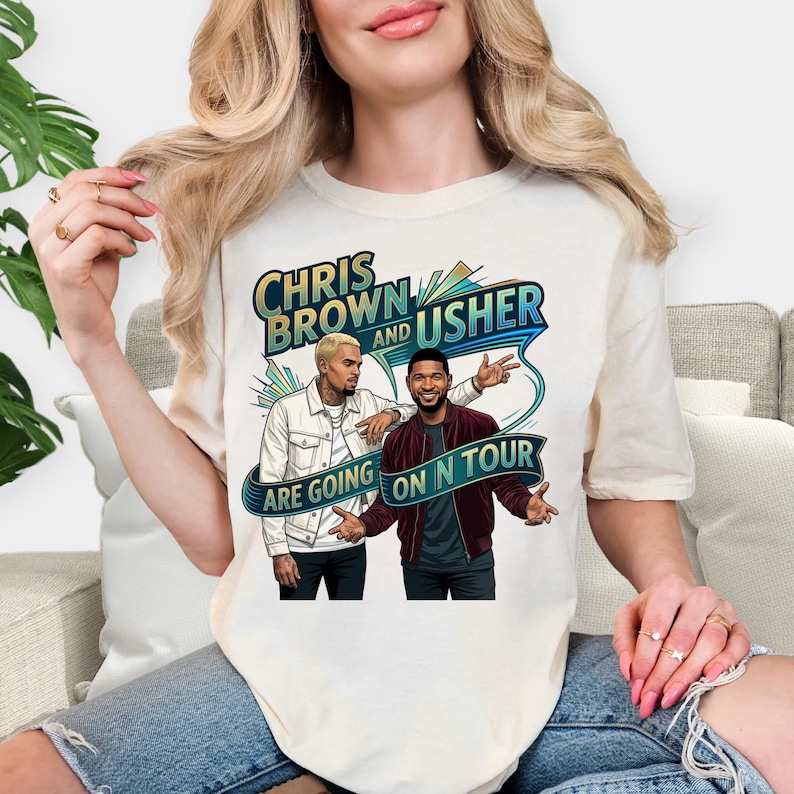 May include: Cream-colored t-shirt featuring a graphic of Chris Brown and Usher. The design includes the text "CHRIS BROWN and USHER ARE GOING ON N TOUR" in a stylized font with blue and green accents. The illustration depicts the two artists.