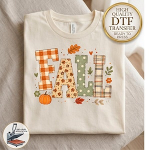May include: A cream-colored t-shirt featuring the word FALL in large block letters. Each letter has a different pattern: orange plaid, floral print, sage green with white dots, and a multi-colored plaid. Includes a pumpkin and leaf graphics.