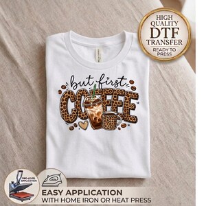 May include: A white t-shirt featuring a graphic design with the text but first COFFEE in a leopard print pattern. The design includes an iced coffee cup and a mug, both with leopard print details, surrounded by scattered coffee beans.