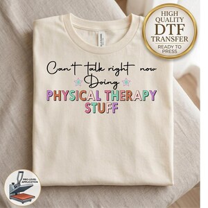 May include: A cream-colored t-shirt featuring the black text "Can't talk right now Doing" above the multi-colored text "PHYSICAL THERAPY STUFF" in a bold font, accented by two small stars with light green centers and pink outlines.