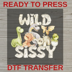 May include: DTF transfer design featuring the text "WILD SISSY" with various cartoon dinosaurs in shades of green, orange, gray, and yellow. The words "READY TO PRESS" and "DTF TRANSFER" are in red.