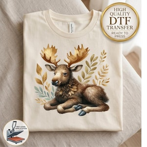 May include: A cream-colored t-shirt featuring a graphic of a brown moose lying down, surrounded by golden-toned leaves. The moose has large, detailed antlers. A circular gold badge in the top right corner reads High Quality DTF Transfer Ready To Press.