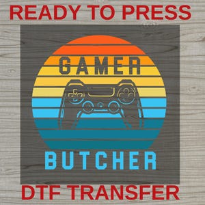 May include: A graphic design featuring a sunset motif with horizontal stripes in orange, yellow, and blue. The text reads "READY TO PRESS" at the top, "GAMER" above a black game controller silhouette, "BUTCHER" below, and "DTF TRANSFER" at the bottom.