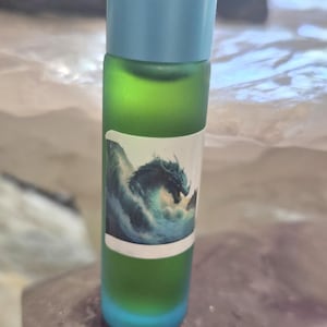 May include: A cylindrical glass roller bottle with a frosted green finish and a light blue cap. A white rectangular label is applied to the front, featuring a detailed illustration of a dark dragon emerging from turbulent blue ocean waves.