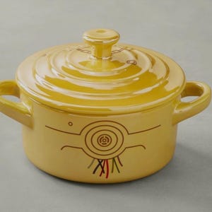 Może przedstawiać: Żółte ceramiczne naczynie do zapiekania z pokrywką. Naczynie ma dwa uchwyty i dekoracyjny wzór z przodu. Wzór zawiera spiralę i linie w kolorze brązowym, czerwonym, zielonym i czarnym.