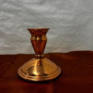 May include: A shiny, copper-colored candlestick holder. The holder has a wide, rounded base, a slender stem, and a small cup at the top for holding a candle. The metal reflects light, creating a warm, inviting glow. The candlestick holder is a decorative item.