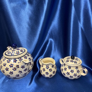 May include: A ceramic tea set with a teapot, creamer, and sugar bowl. The white pieces are decorated with blue and orange floral patterns. The teapot has a lid and spout. The sugar bowl has a lid and handles. The creamer has a handle.