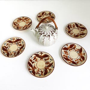 May include: A white ceramic teapot with a woven handle and a matching lid, surrounded by six small saucers. The saucers and teapot feature a brown and cream-colored bird design with blue accents. The set is arranged on a white surface.