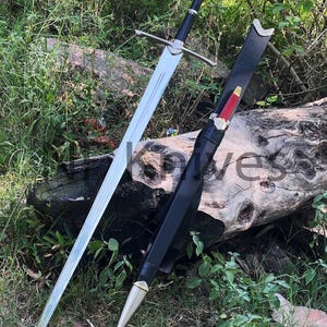 May include: A long steel sword with a straight double-edged blade, a silver crossguard, and a black grip with a silver pommel. Beside it lies a black leather scabbard featuring a red handle section, silver metal fittings, and a pointed silver tip.