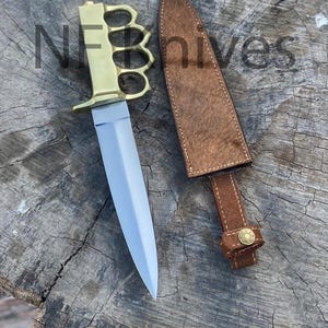 Könnte beinhalten: Ein zweischneidiger Dolch mit einer polierten silbernen Klinge und einem Schlagringgriff aus Messing. Daneben liegt eine braune, strukturierte Lederscheide mit einem Druckknopf aus Messing. Der Text NFknives ist im Bild zu sehen.