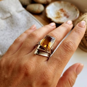 May include: A silver ring featuring a large, rectangular, faceted amber-coloured gemstone set in a polished bezel. The band consists of multiple layered, textured silver strips that wrap around the finger, creating a wide, architectural design.