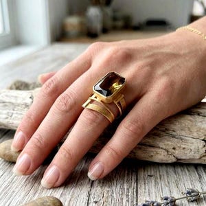 May include: A gold ring featuring a large, rectangular, faceted dark brown gemstone set in a polished metal frame. The band consists of multiple thin, layered gold wires with a textured finish that wrap around the finger in a wide design.