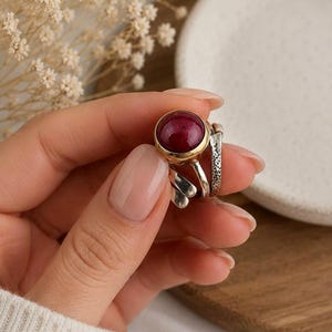 May include: A ring featuring a round, deep red cabochon gemstone set in a gold-toned bezel. The band is composed of multiple silver-toned segments, including one with a textured, hammered finish and others with smooth, polished surfaces.