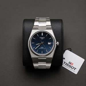 Op de afbeelding: Een Tissot PRX Powermatic 80 polshorloge met een geïntegreerde zilverkleurige roestvrijstalen band en een getextureerde donkerblauwe wijzerplaat. De wijzerplaat heeft zilveren indexen, een datumvenster op de drie-uurpositie en een wit Tissot-label.