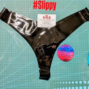 Op de afbeelding: Zwarte latex string. Het ondergoed is glanzend en heeft een V-vormig ontwerp. Het woord "#Slippy" staat in het rood bovenaan. Een kleine kaart en een ronde sticker zijn ook zichtbaar.