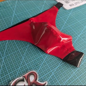 Könnte beinhalten: Eine rote Latex-Gesichtsmaske mit schwarzem Besatz. Die Maske hat eine konturierte Form und eine glänzende Oberfläche. Sie wird auf einer grünen Schneidematte gezeigt.
