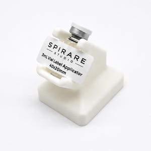 May include: A white plastic 3mL vial label applicator with a square base and a cradle for holding a small glass vial. The device features a label with the text SPIRARE STUDIO, 3mL Vial Label Applicator, and 40x20mm printed in black ink.