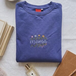 Könnte beinhalten: Ein periwinkelfarbenes Sweatshirt mit dem gestickten Wort "Mama", umgeben von bunten Blumen und Blättern. Das Sweatshirt ist auf einer weißen Oberfläche gefaltet, mit einer Parfümflasche, einem Notizbuch und einer Schale mit Schmuck in der Nähe.