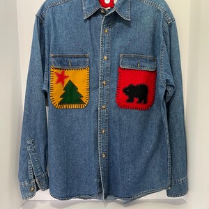 May include: A blue denim long-sleeve button-down shirt featuring two chest pockets with decorative felt patches. The left pocket displays a green pine tree and red star, while the right pocket features a black bear silhouette on a red background.