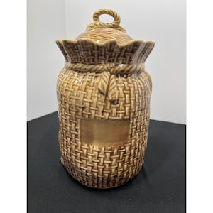 May include: A tan ceramic canister designed to resemble a tied burlap sack. The container features a textured woven pattern, a sculpted rope tie around the neck, and a matching lid with a small loop handle on top. A smooth rectangular area is visible.