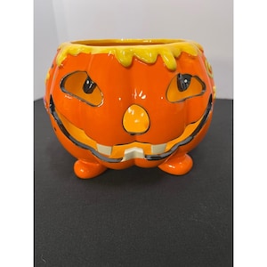 May include: A bright orange ceramic jack-o'-lantern bowl featuring a glossy finish, a wide grinning mouth with visible teeth, triangular eyes, and a rounded nose. The top rim is decorated with a yellow, dripping glaze effect, and the bowl stands on three small, rounded orange feet.