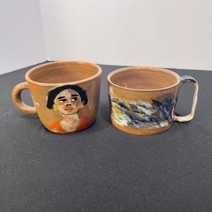 May include: Two ceramic mugs with a glossy tan finish. The left mug features a hand-painted portrait of a person with dark hair and an orange shirt. The right mug displays an abstract pattern of blue, yellow, and white brushstrokes on the side.