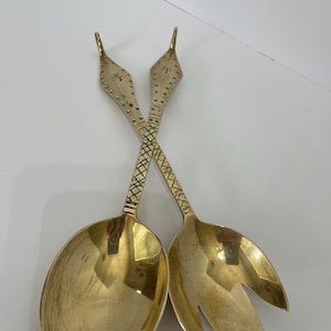 May include: A pair of gold-colored metal salad servers with long, slender handles featuring a diamond-shaped top and a cross-hatched pattern. One server has a solid oval bowl, while the other has a three-pronged fork design at the end.