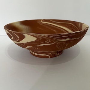 May include: A shallow bowl featuring a marbled pattern of deep terracotta brown and creamy off-white swirls. The bowl has a wide, open circular rim and sits on a raised, pedestal-style circular base, showcasing a continuous swirled design.