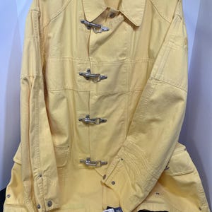 May include: A bright yellow long-sleeve jacket featuring a classic collar and four silver-toned metal firemen-style hook-and-loop closures down the front. The garment includes a Polo Ralph Lauren brand label at the neck and a small tag attached.