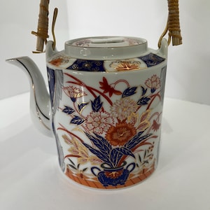 May include: A white porcelain teapot featuring a vibrant floral pattern in shades of deep blue, orange, and gold. The teapot has a curved spout, a matching lid, and a curved handle wrapped in light brown woven rattan material.