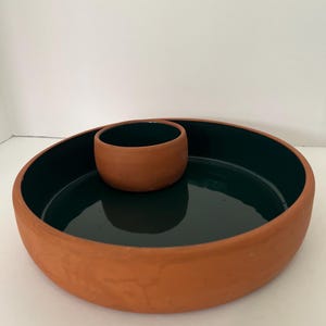 May include: A round, shallow terracotta serving dish with a smooth, dark forest green glazed interior. A smaller, matching round terracotta bowl with a dark green glazed interior is positioned inside the larger dish, creating a two-piece set.