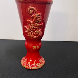 May include: A bright red ceramic goblet featuring a raised, embossed scrollwork design on the front. The rim, the base, and the central stem area exhibit a distressed, weathered finish with patches of exposed tan texture against the red glaze.