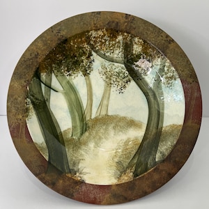 May include: A round ceramic bowl featuring a hand-painted forest scene with tall green trees, a grassy hill, and a light blue sky. The wide rim has a textured, mottled brown and reddish-brown finish that frames the central landscape illustration.