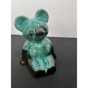 May include: A ceramic bear figurine featuring a glossy, mottled teal and black glaze. The bear is depicted in a seated position with rounded ears, a defined snout, and a smooth, reflective surface finish that highlights the color variation.