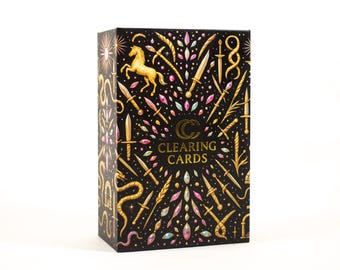 May include: A rectangular deck box for Clearing Cards featuring a black background with gold illustrations of a horse, swords, snakes, and wheat, accented by small pink and blue gems. The center displays the text Clearing Cards in gold.