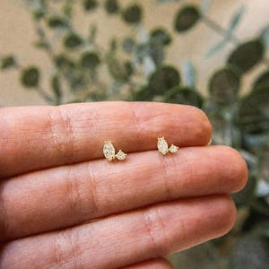 May include: Two gold stud earrings, each featuring a marquise-cut clear gemstone paired with a smaller round-cut clear gemstone. The stones are set in a gold metal base, creating a delicate, asymmetrical cluster design for each individual earring.
